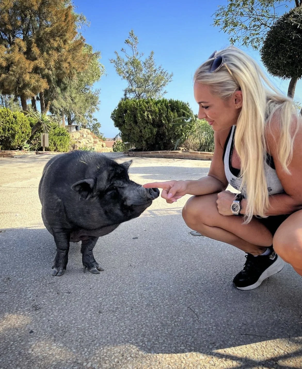 A woman with long blonde hair crouches and interacts with a black pig outdoors on a paved area, surrounded by greenery and trees.