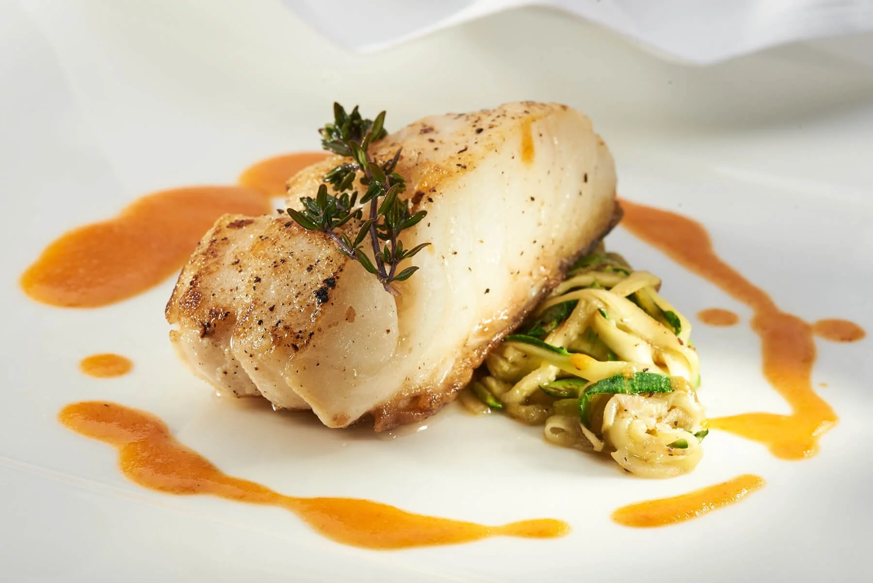 Gebratenes Fischfilet auf einem weißen Teller, garniert mit einem Zweig Kräuter, dazu eine Nudelsalat und eine orangene Sauce.