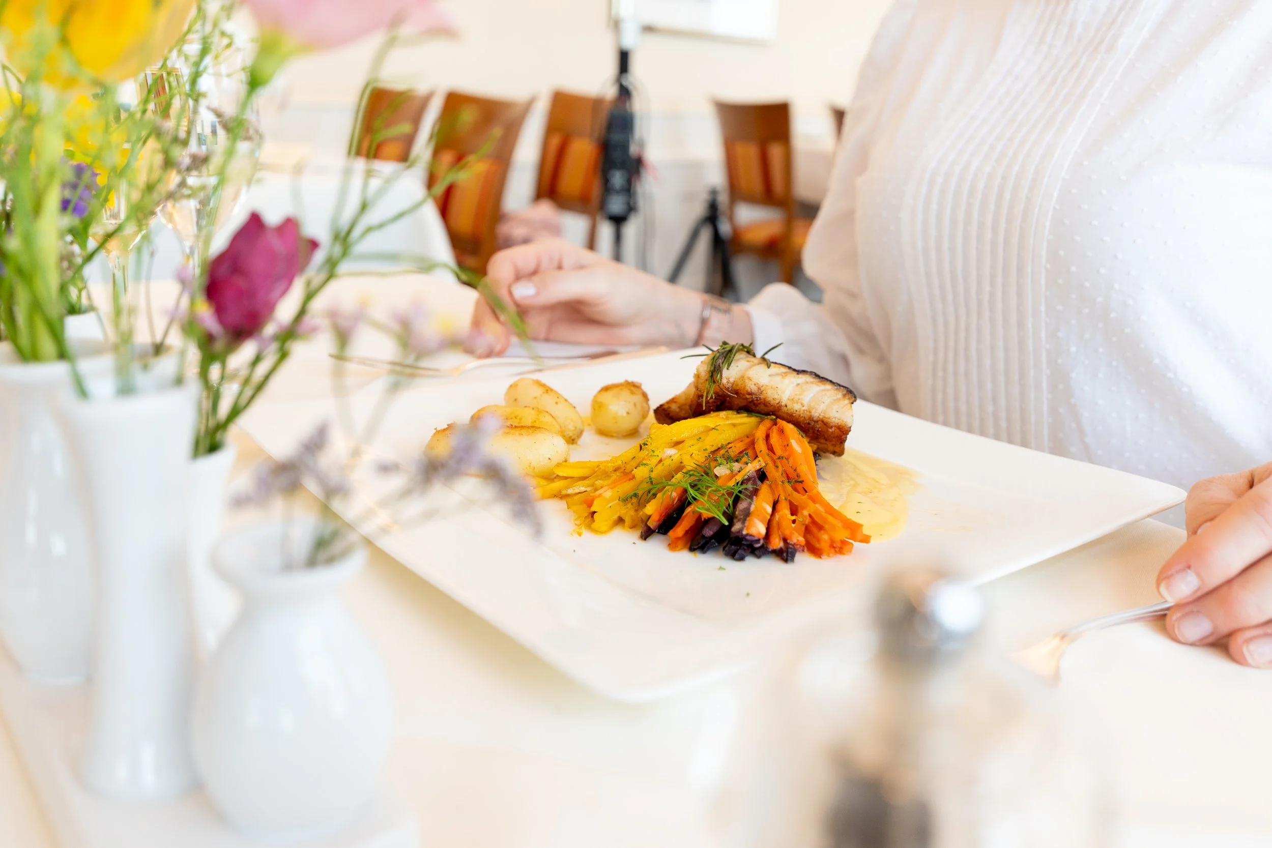 Ein Teller mit einem Gericht aus Fisch, grünen Sprossen und Sauce, umgeben von Weingläsern, Wasserflaschen und Brot auf einem festlich gedeckten Tisch.