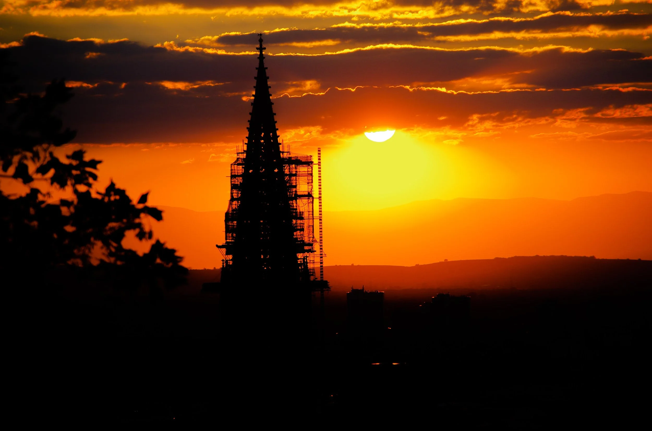 Sonnenuntergang hinter dem Kirchturm mit Baugerüsten, Silhouetten von Gebäuden im Vordergrund, bewölkter Himmel, warme Farbtöne.