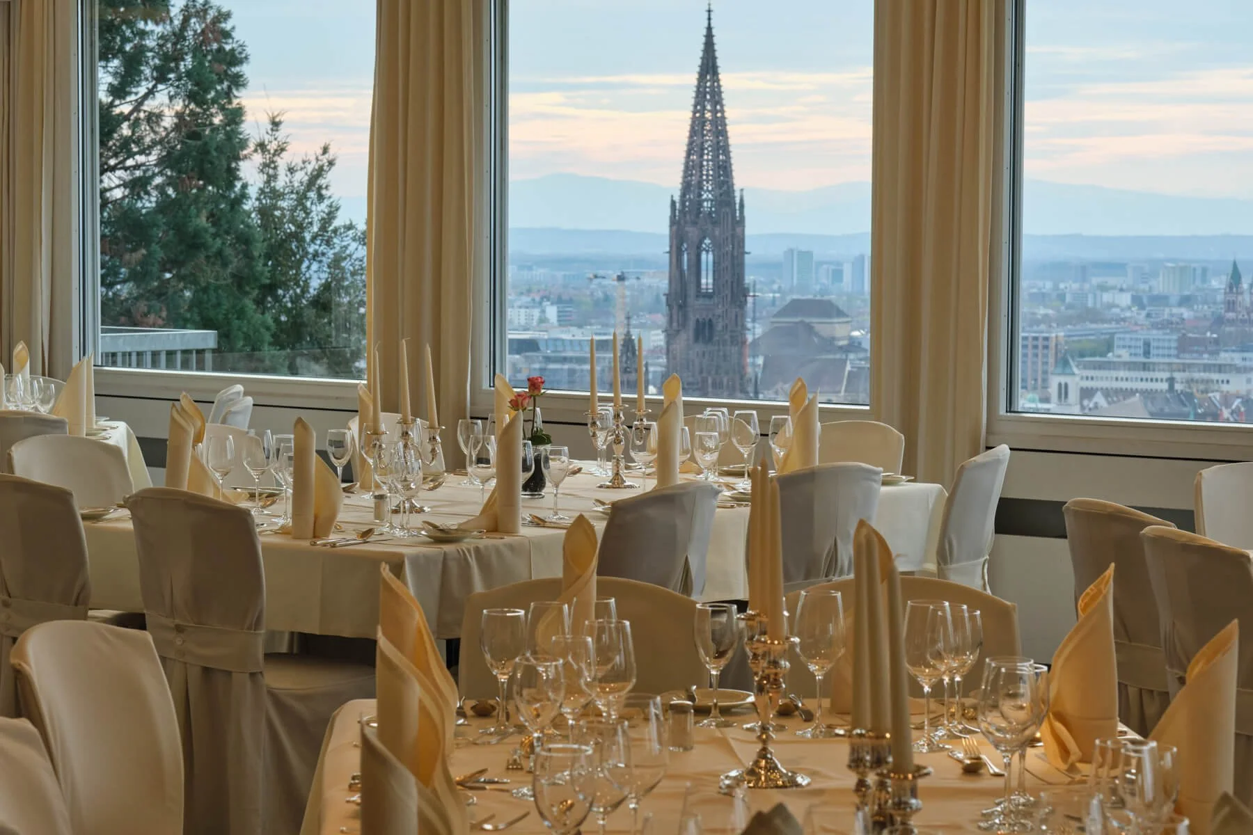 Ein festlich gedeckter Speisesaal mit mehreren Tischen, weißen Stoffdecken, Kerzenhaltern und Weingläsern, Blick auf eine Stadtkathedrale durch große Fenster, bei Tageslicht.