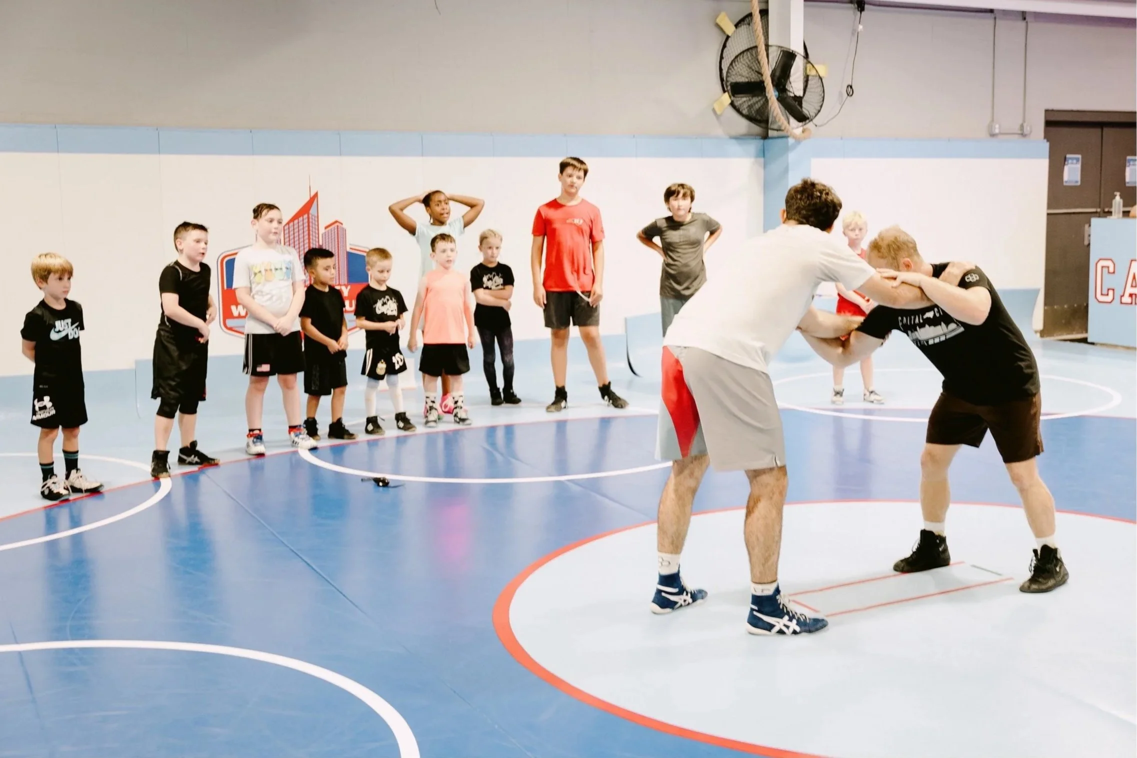 A group of children watching two adults sparring in a gym. The children stand in a line, some with hands on their heads, wearing athletic clothes. The sparring adults are engaged in a wrestling move, on a blue and orange basketball court. There are posters and a fan on the wall in the background.