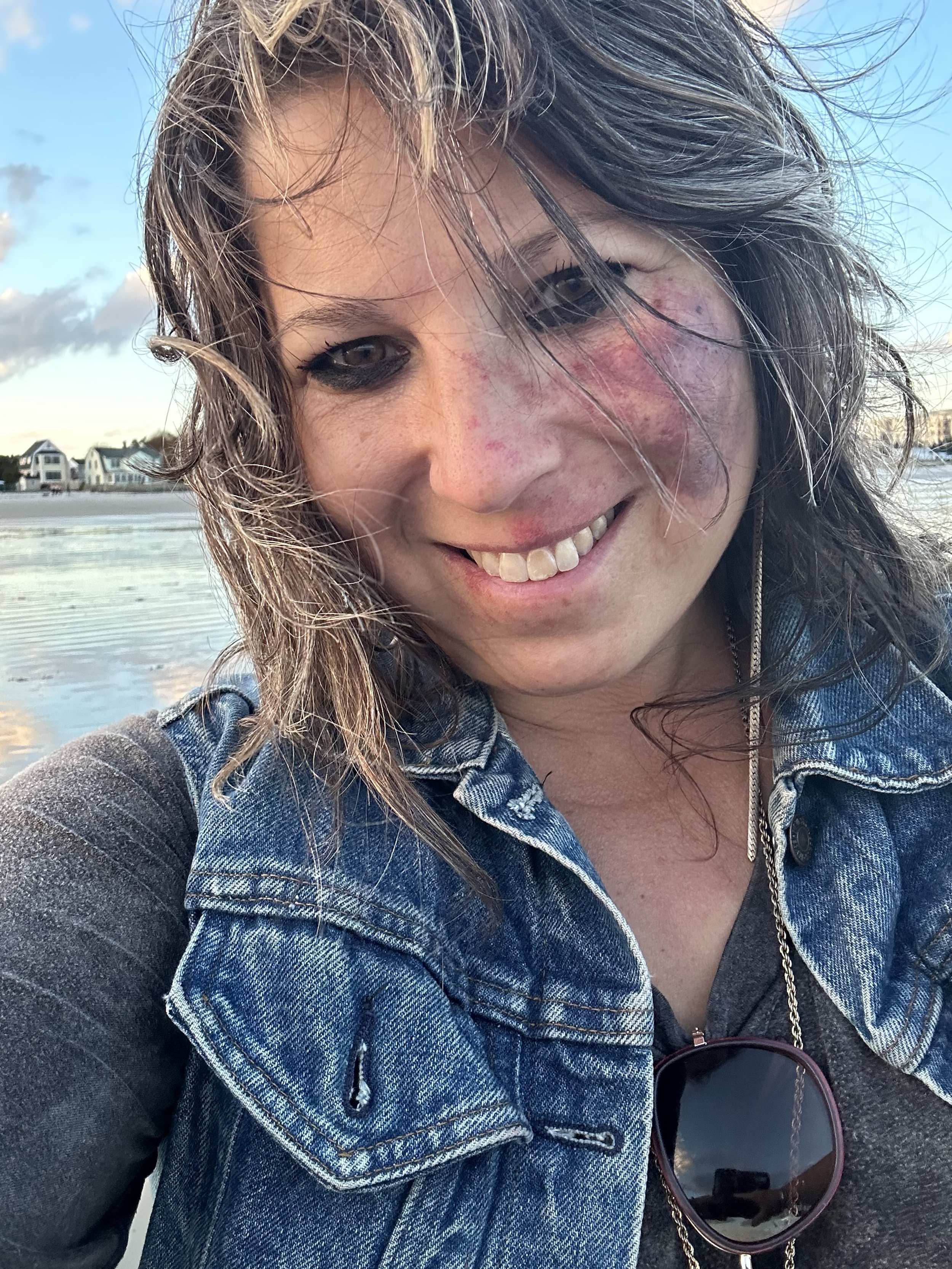 Person smiling at the camera wearing a denim vest with sunglasses hanging from the collar, standing on a beach with houses in the background.