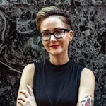 Person with short hair and glasses wearing a sleeveless black top standing against a textured wall.