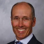 Smiling man wearing glasses and a suit with a gray background.