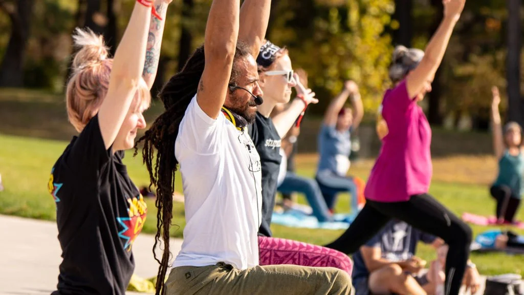 Group of people practicing yoga outdoors with raised arms, led by an instructor with a microphone.