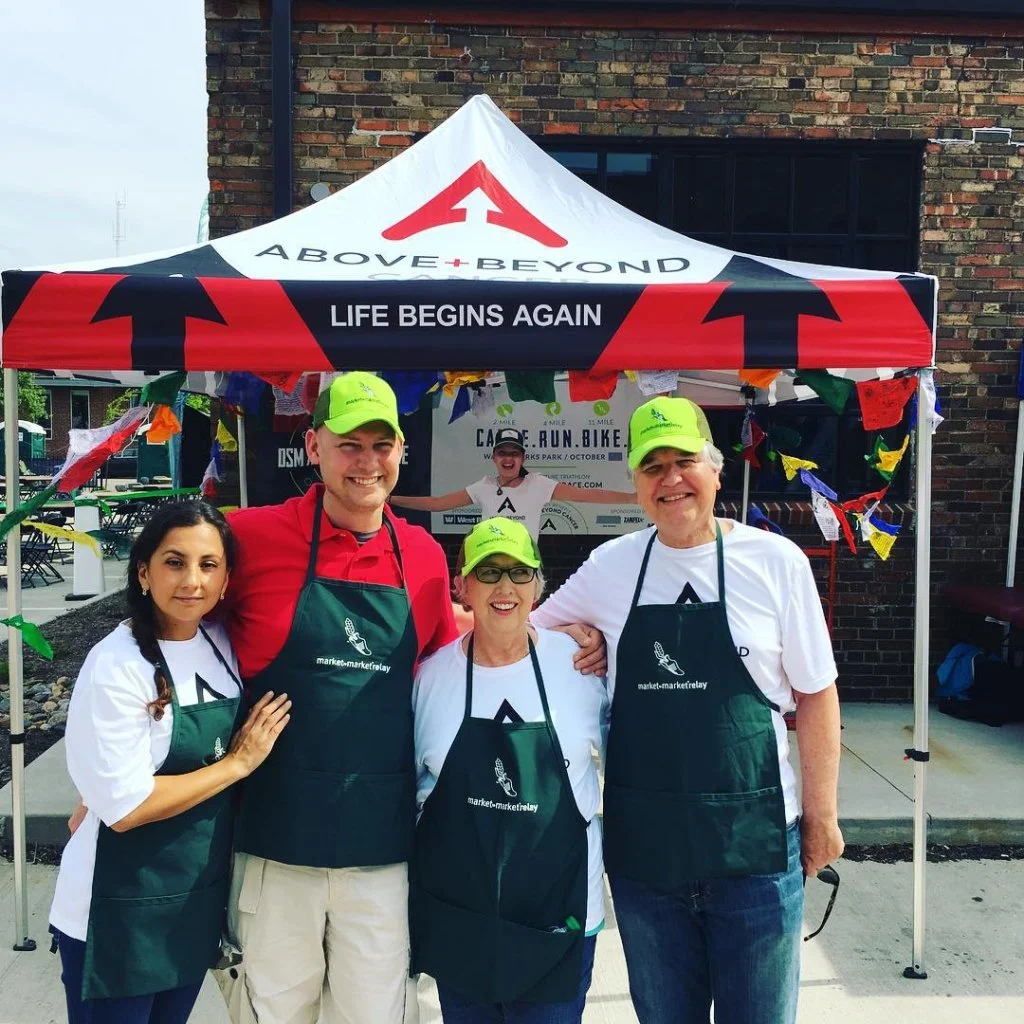 Four people standing under a canopy with 'Above + Beyond' and 'Life Begins Again' printed on it. They are wearing aprons with a market symbol, and green hats. A banner in the background promotes a 'Bike Run' event.