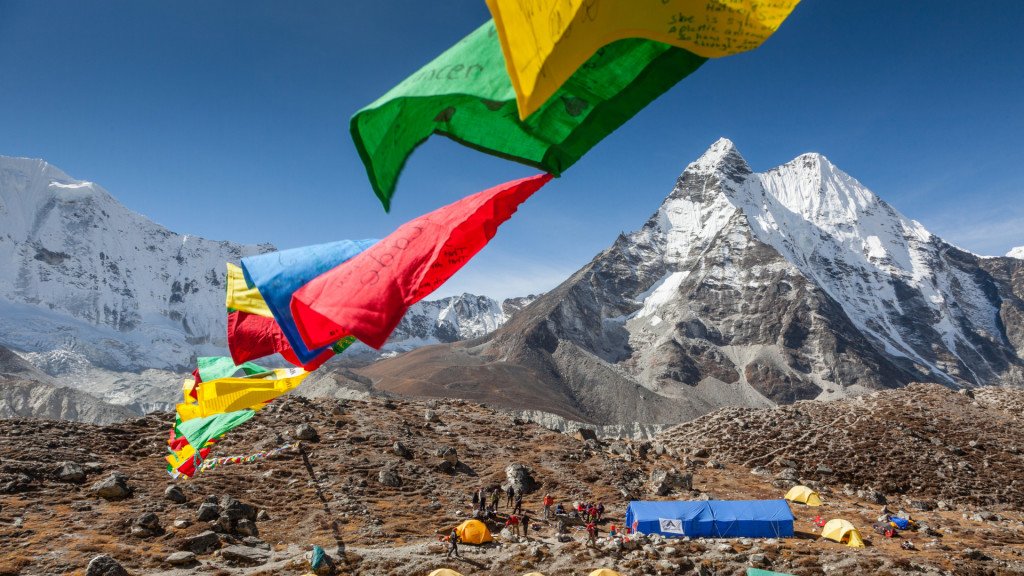 Himalayan mountains with snow, colorful prayer flags in the foreground, and a campsite with tents.