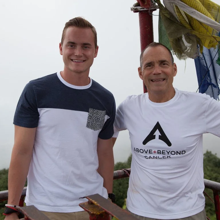 Two men smiling, one wearing a blue and white shirt and the other a shirt with "Above + Beyond Cancer" logo, standing outdoors near flags.