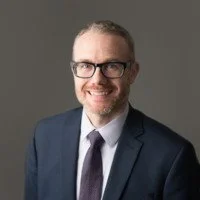 Man wearing glasses and a suit smiling against a gray background.