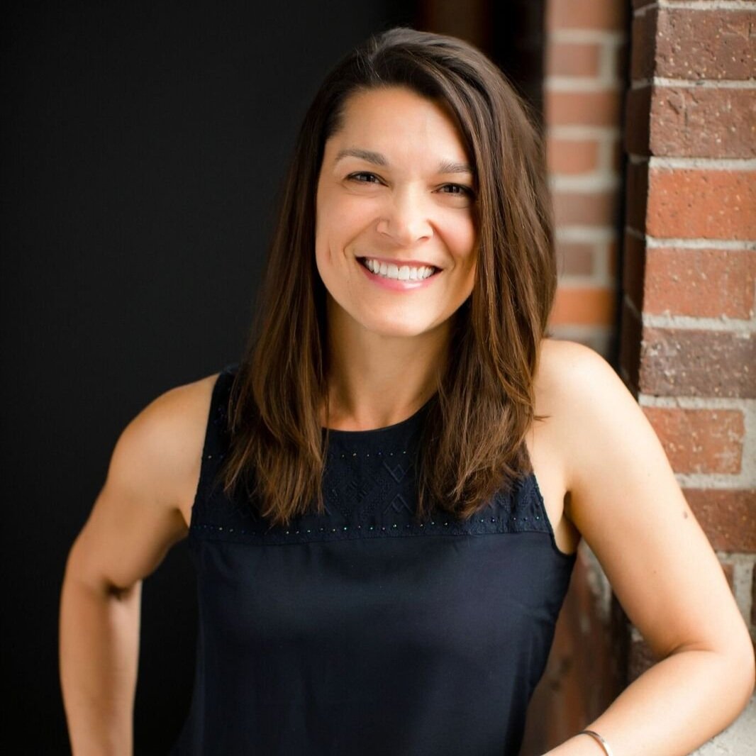 Smiling woman in a sleeveless black top leaning against a brick wall.