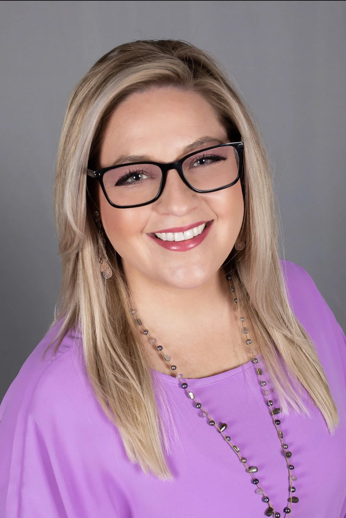 Smiling person with glasses and blonde hair wearing a purple top and beaded necklace.