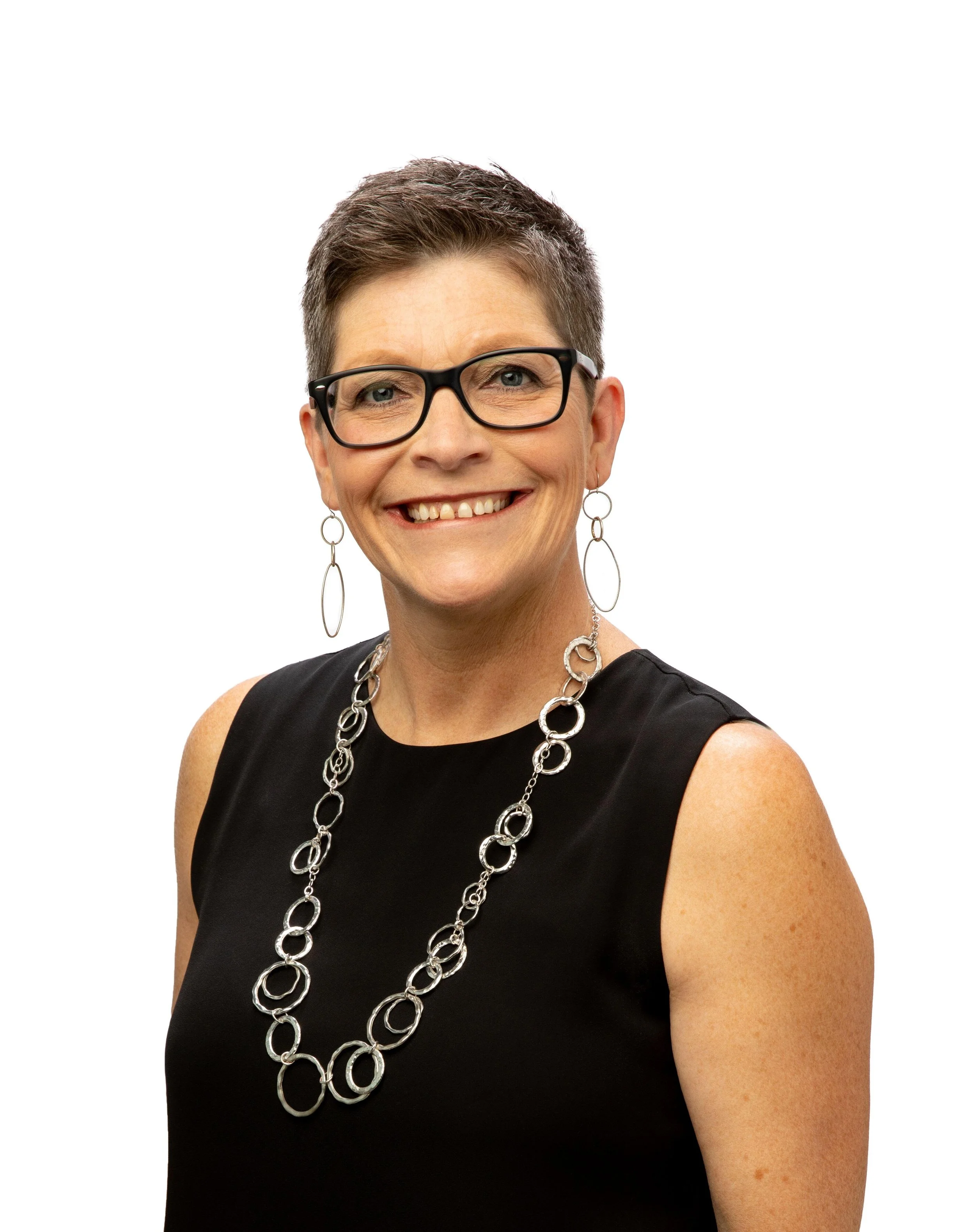 Smiling person with short hair, glasses, hoop earrings, and a necklace, wearing a sleeveless black top, on a white background.