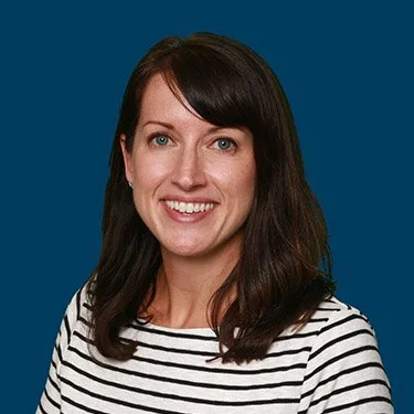 A woman smiling, wearing a black and white striped shirt, in front of a solid blue background.