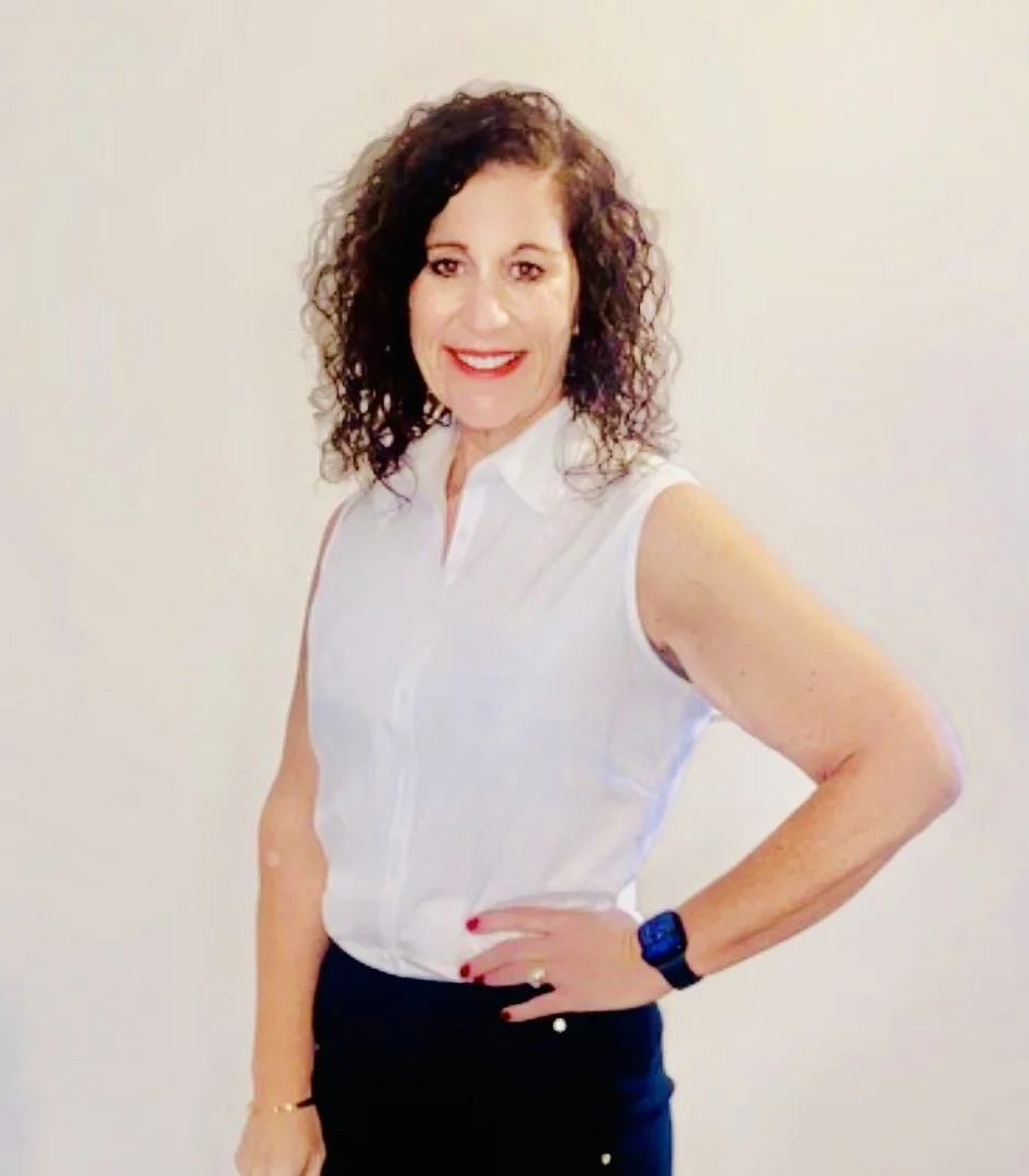 Smiling woman with curly hair wearing a sleeveless white blouse and black pants, standing against a plain background.