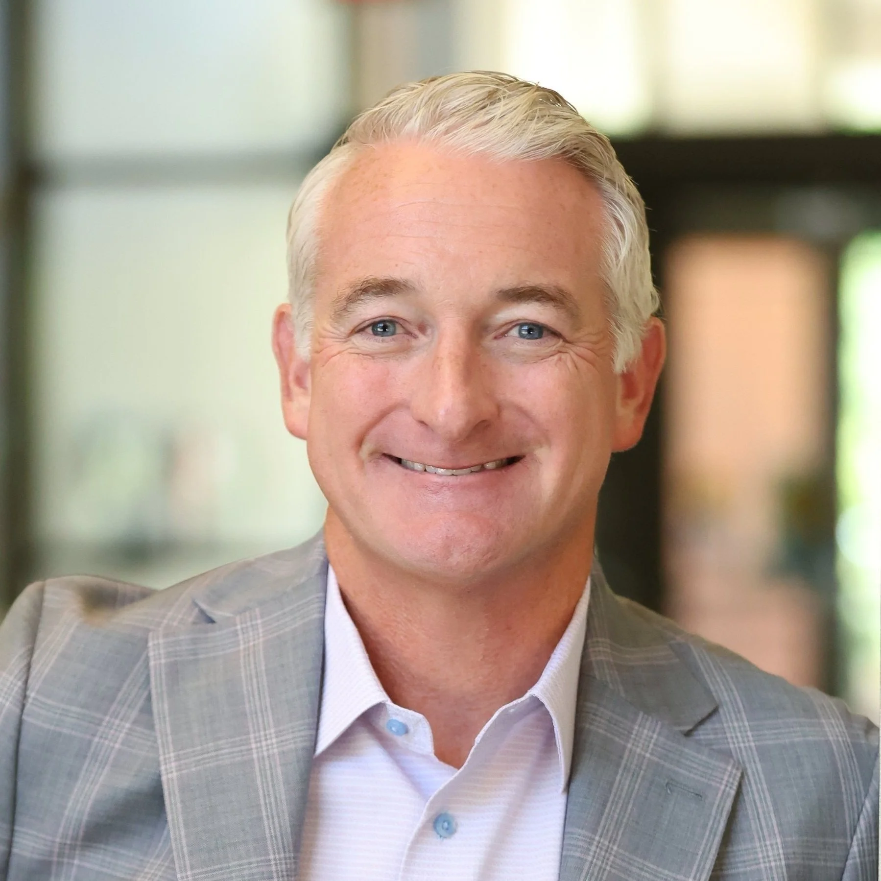 Smiling man in a light gray plaid suit jacket with a light-colored shirt, indoors with blurred background.