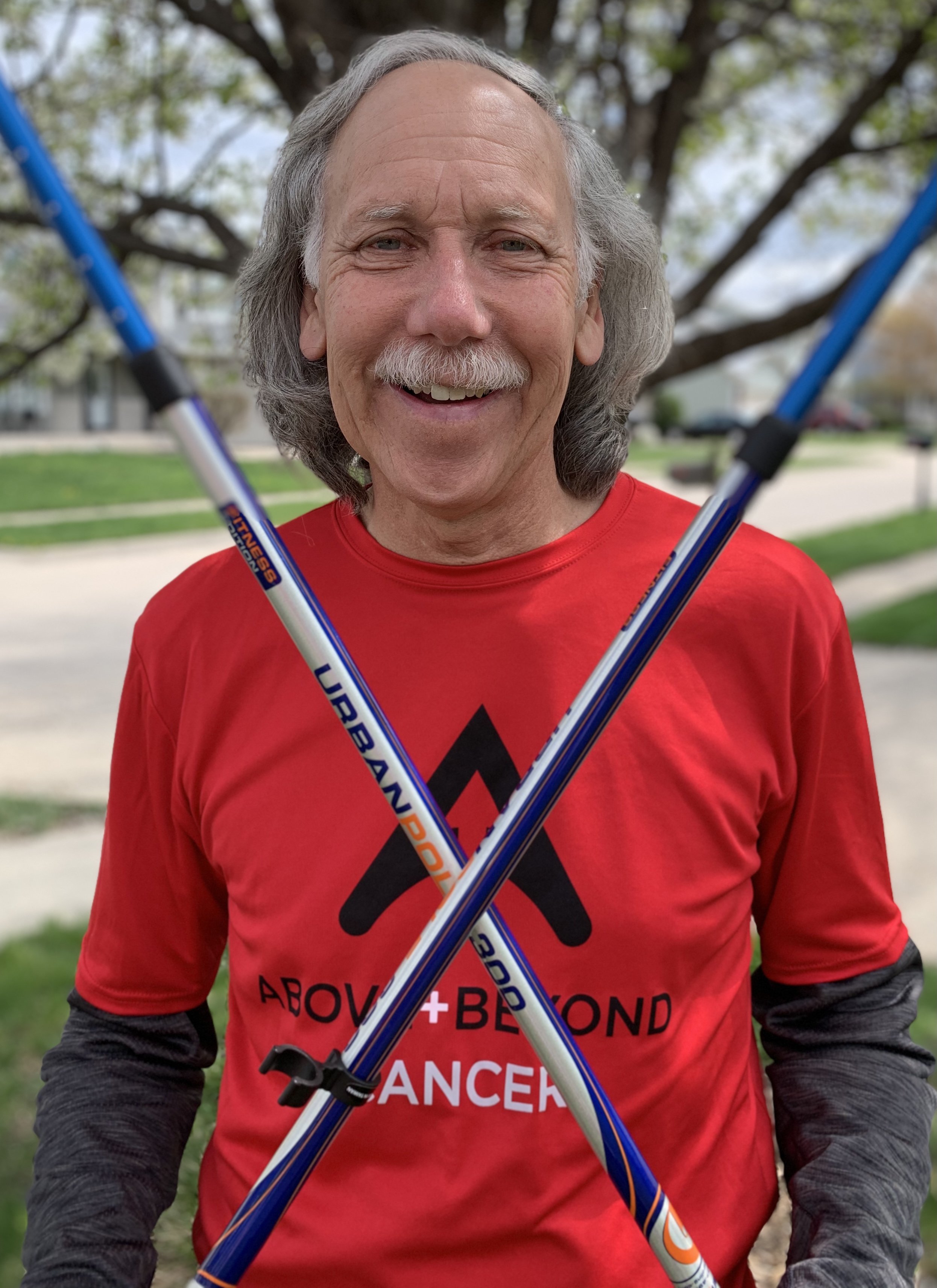Man in red shirt smiling, holding crossed walking poles, standing outdoors.