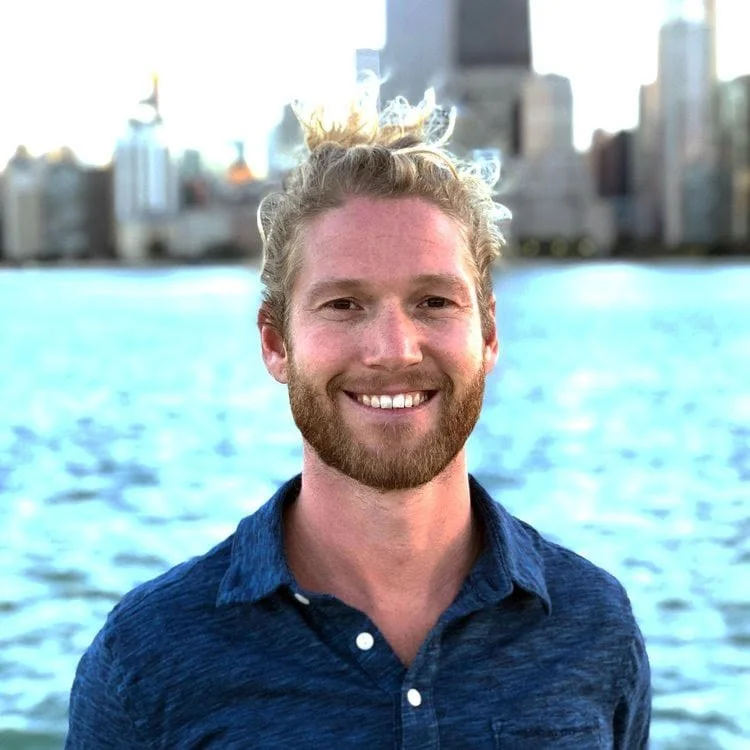 Man smiling in front of a body of water with a city skyline in the background.