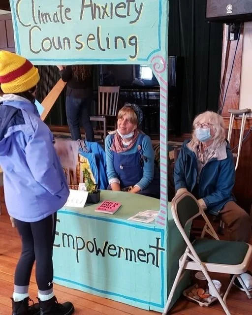 Two older white women sitting at their version of the Climate Anxiety Counseling booth. Signs say "Climate Anxiety Counseling" and "Empowerment." A third person is standing and talking with them, their back to the camera.
