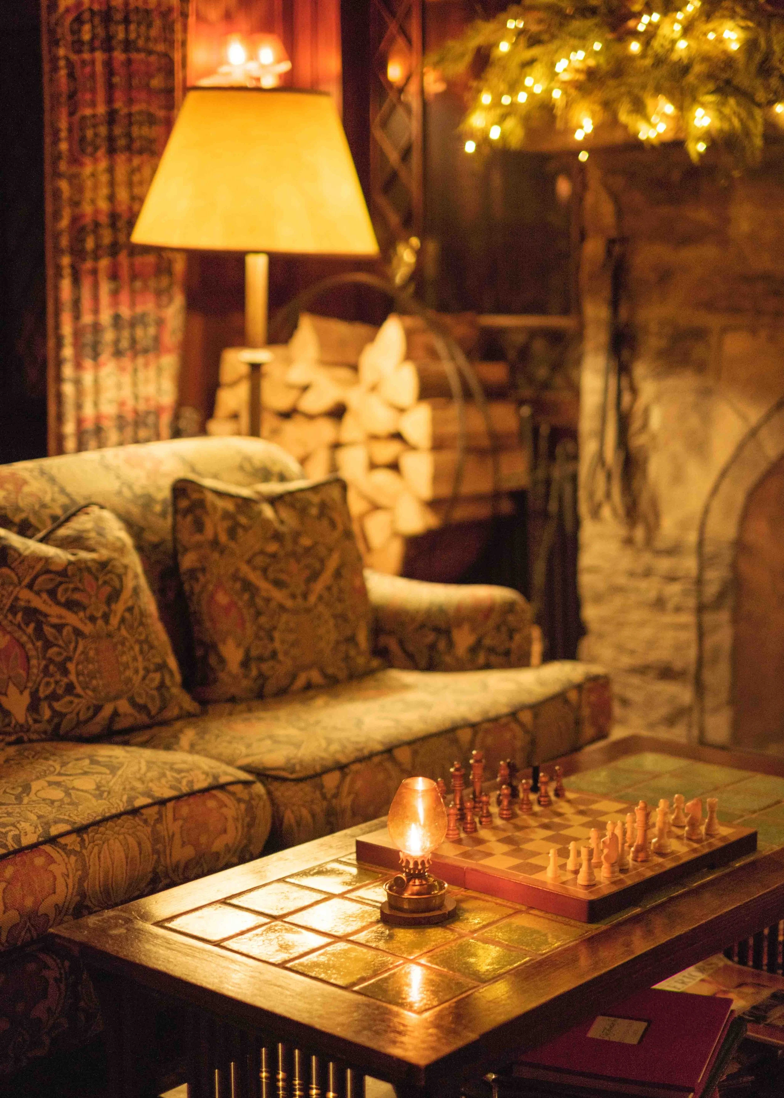 A cozy, dimly lit living room with a wooden coffee table set for chess, a patterned sofa with throw pillows, a table lamp, a stack of firewood, and a lit decorative candle, with a fireplace and Christmas decorations in the background.