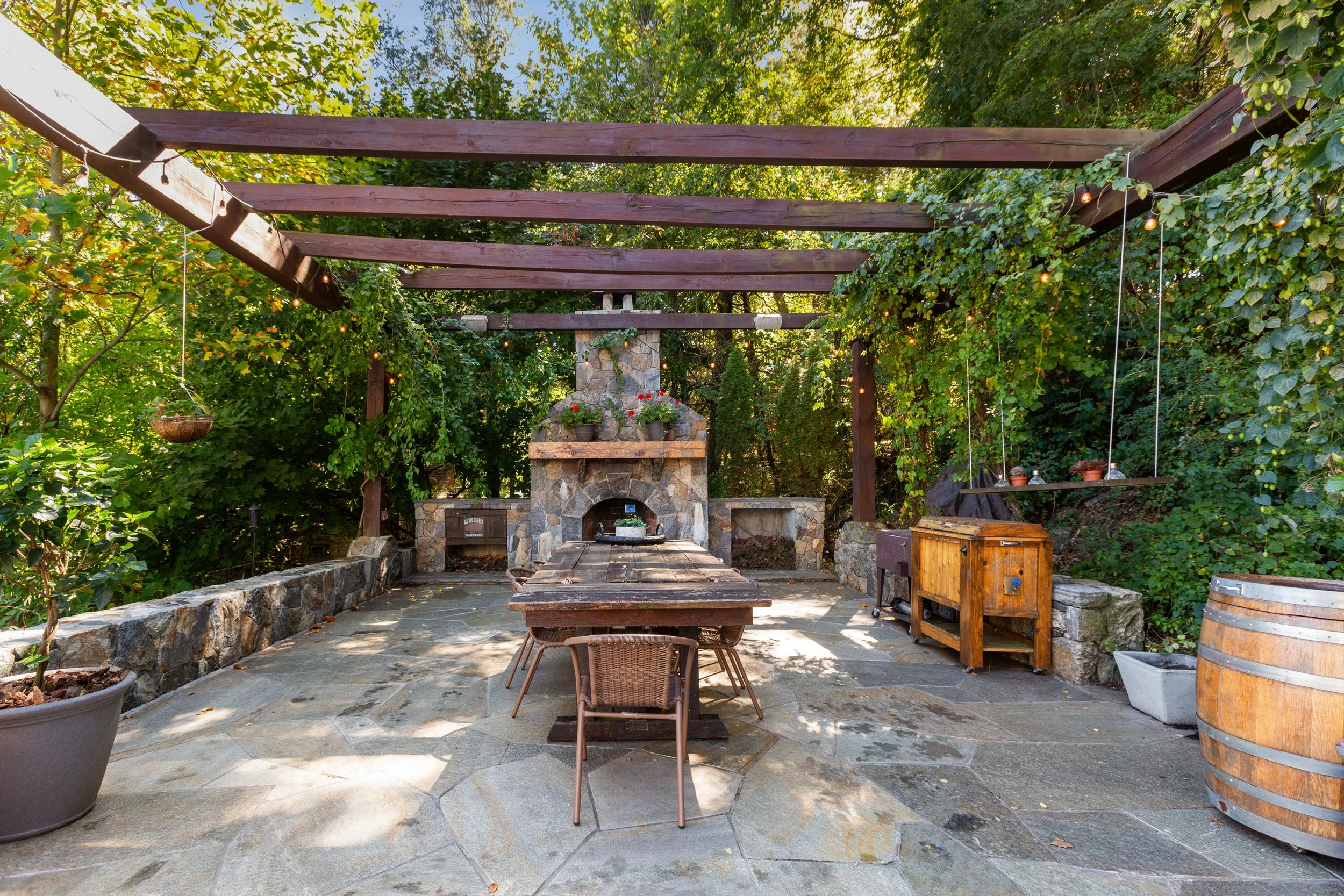 Outdoor patio with stone flooring, wooden table, chairs, a stone fireplace, surrounded by trees and greenery, with string lights overhead.