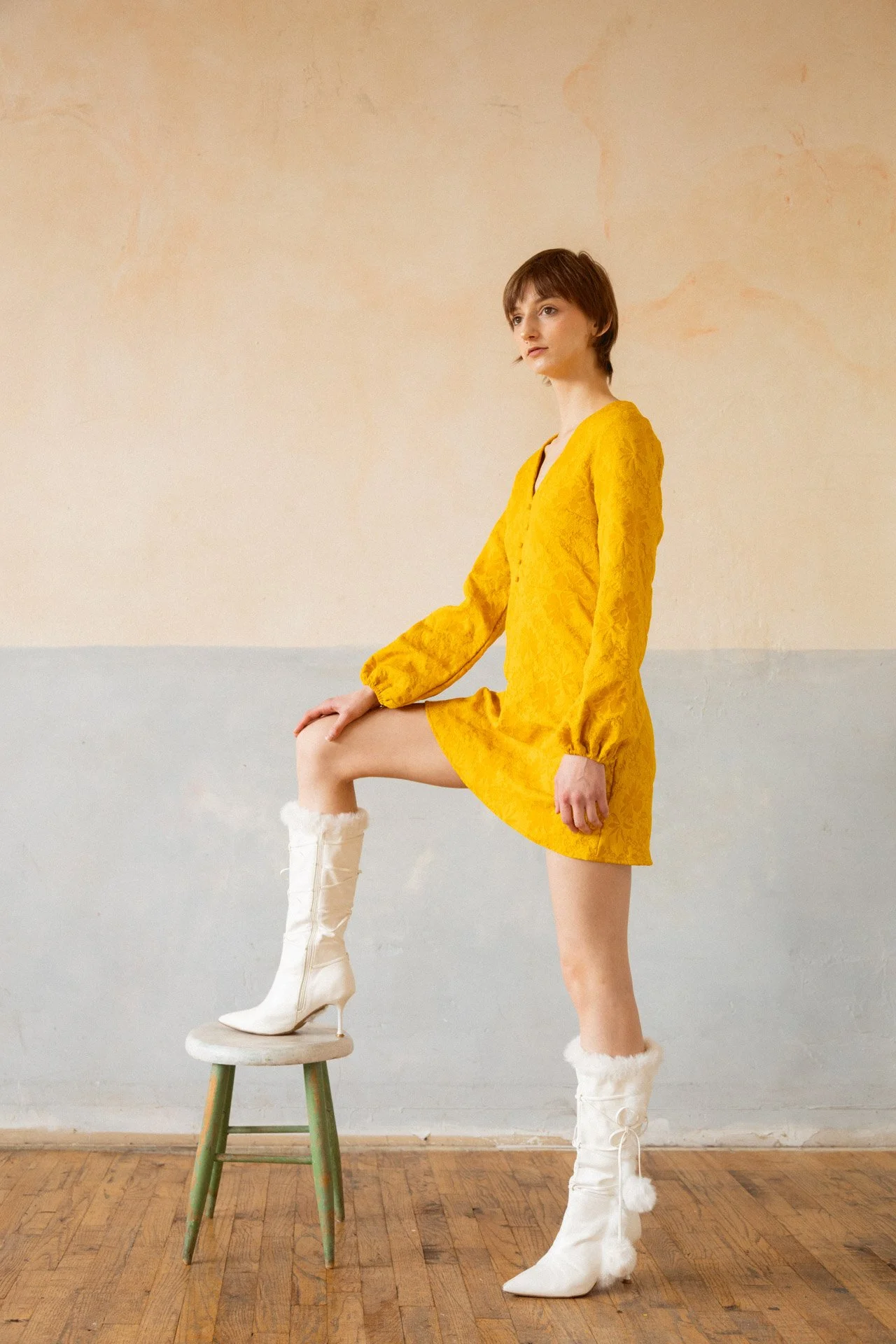 A woman in a yellow dress and white fur-lined boots, with one foot on a stool, standing in a room with a wooden floor and beige wall.