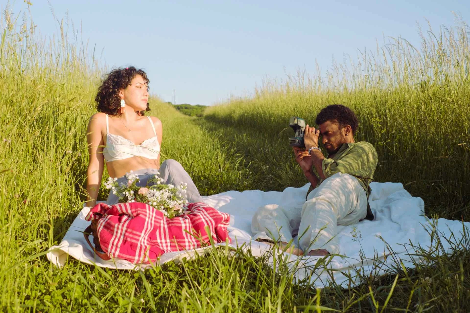 A woman and a man having a picnic in a grassy field with tall green grass on either side. The woman is sitting on a white blanket with a bundle of flowers and a red and white checkered cloth in front of her, wearing a white crop top and light-colored pants. The man is lying on his side, taking a photo of the woman with a camera, dressed in a green shirt and light pants.