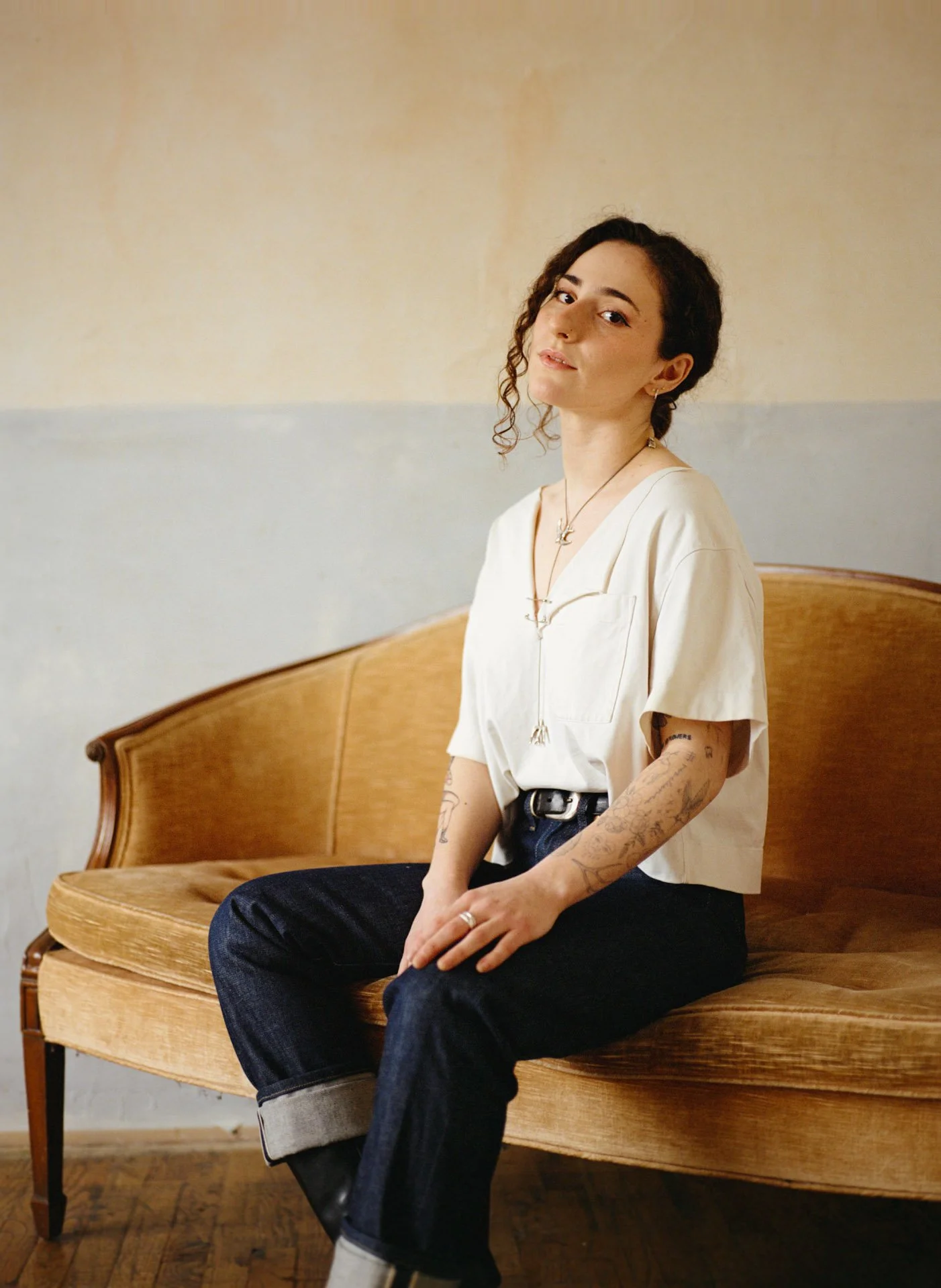 A young woman with curly brown hair and tattoos, sitting on a vintage wooden sofa with a tan fabric cushion, wearing a cream-colored short-sleeve shirt, dark jeans, and white shoes, in a room with a two-toned beige and gray wall.