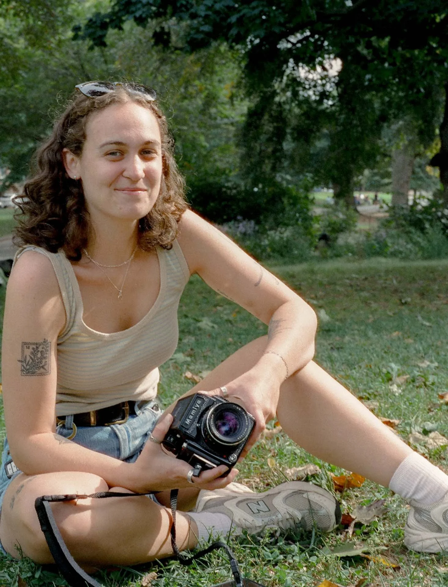 A young woman with shoulder-length curly hair, sitting on grass in a park, holding a vintage camera, smiling at the camera, wearing a beige tank top, denim shorts, white socks, and sneakers, with sunglasses on her head and tattoos on her arm and leg.