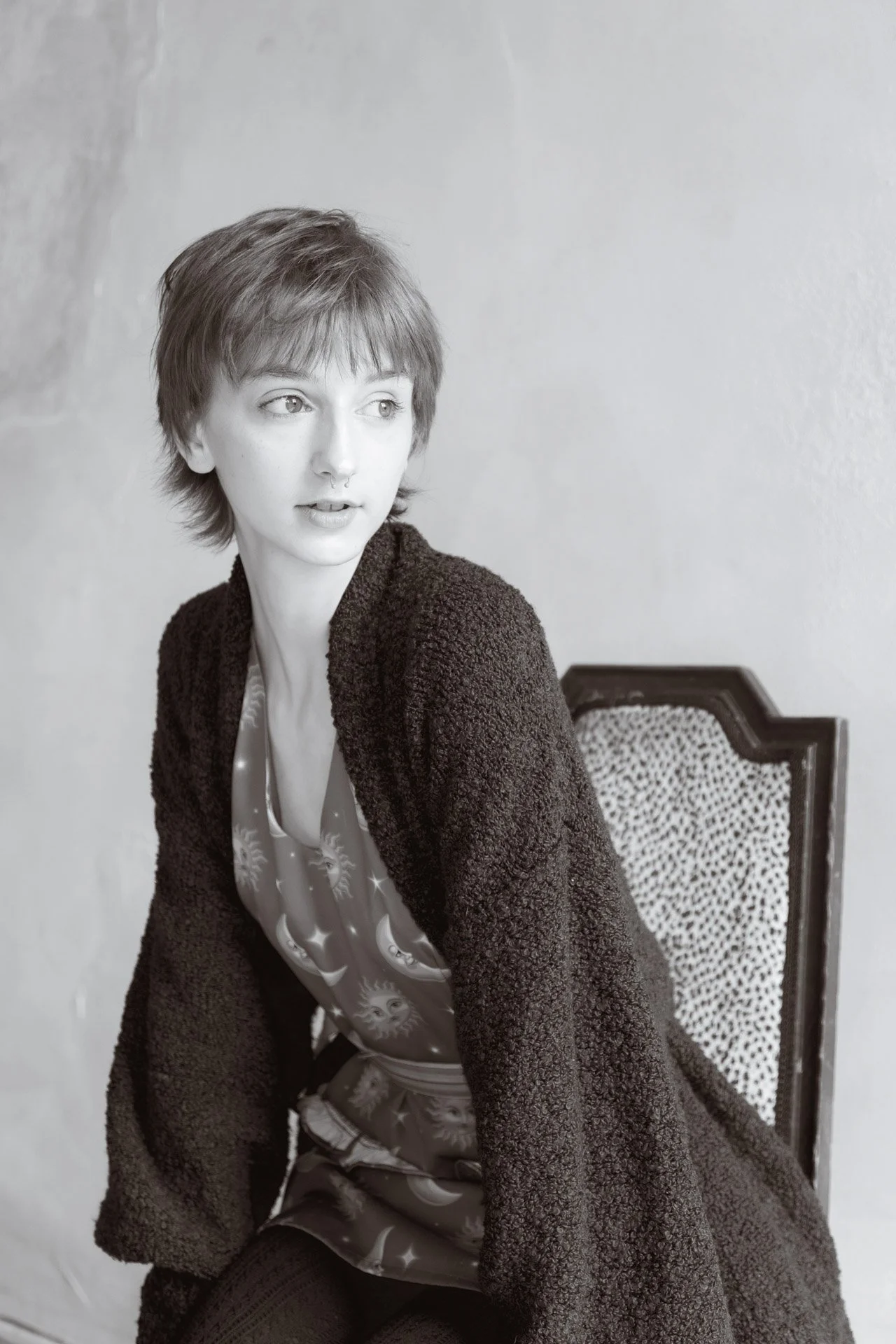 Black and white photo of a young woman with short, tousled hair sitting on a patterned chair, wearing a patterned dress and a fluffy dark cardigan, looking to her left.