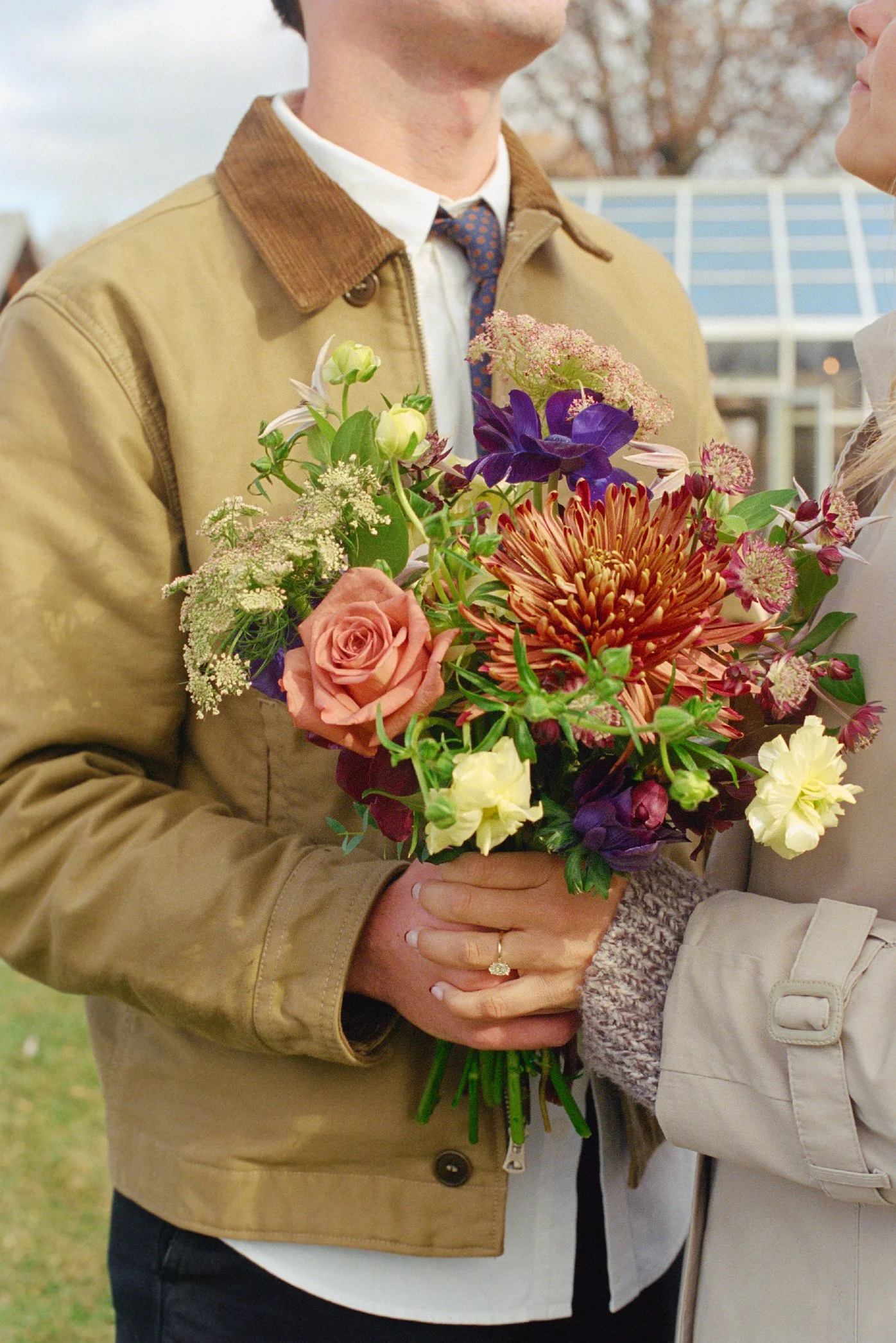 A person in a beige jacket holding a colorful bouquet of flowers, with another person partially visible on the right.