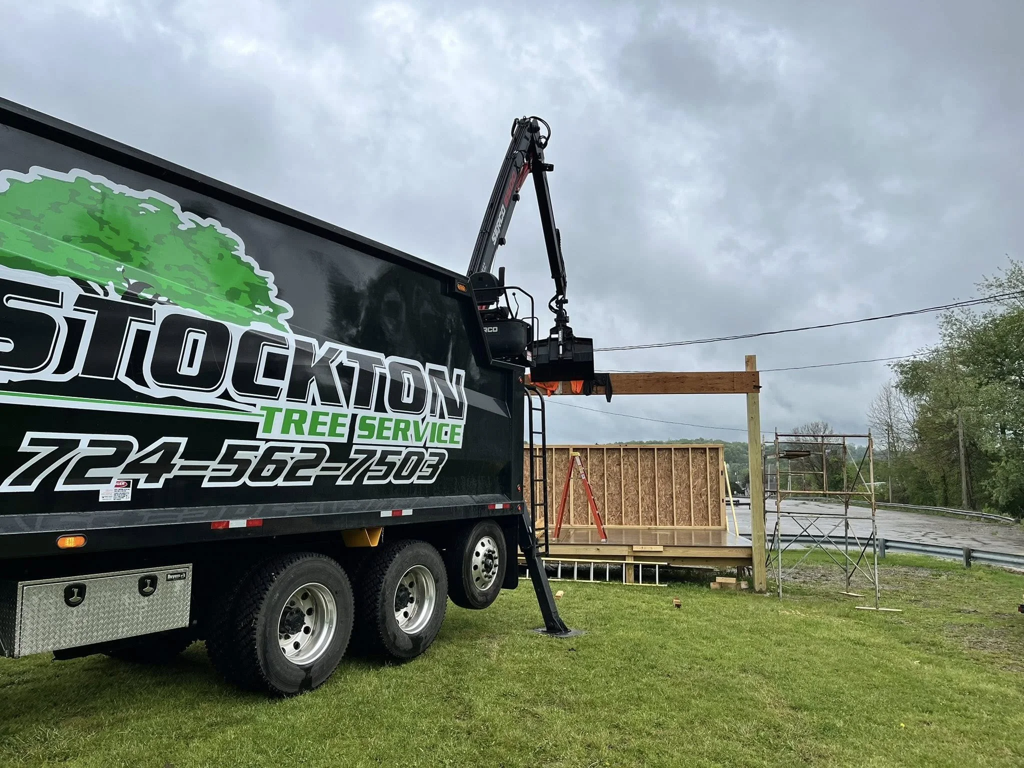 Stockton Tree Service grapple truck parked on grass, with a cherry picker lift extended towards a partially constructed wooden frame for a building, under a cloudy sky.