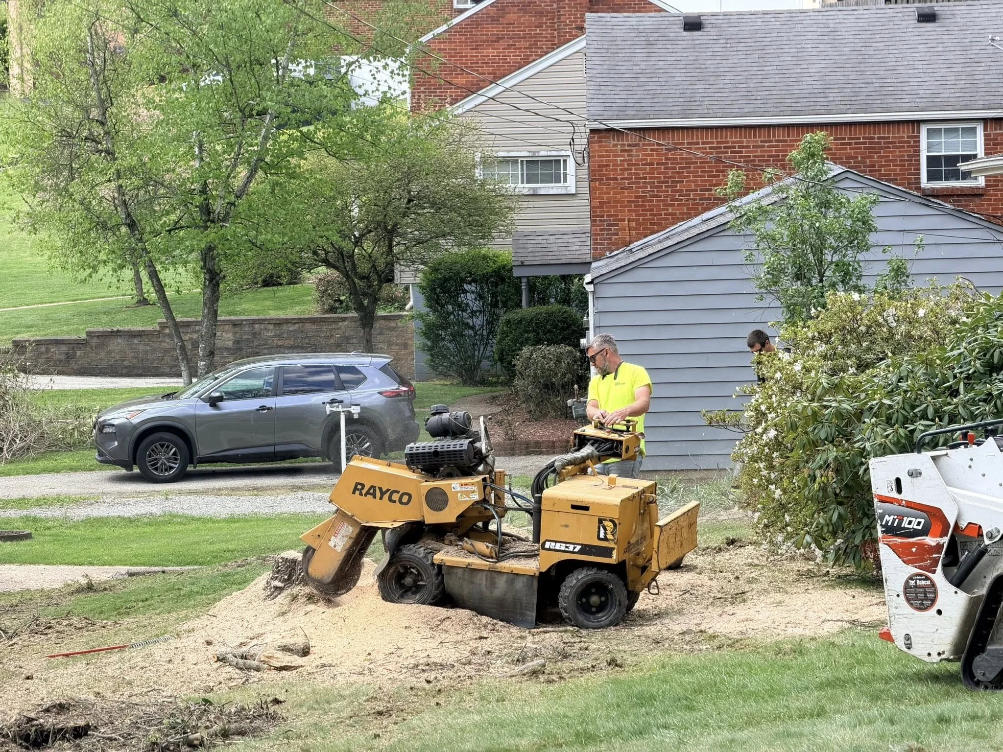 A yellow stump grinder with black and yellow warning labels is grinding a tree stump in a grassy yard near a residential neighborhood. The background features green trees, a street, and houses.