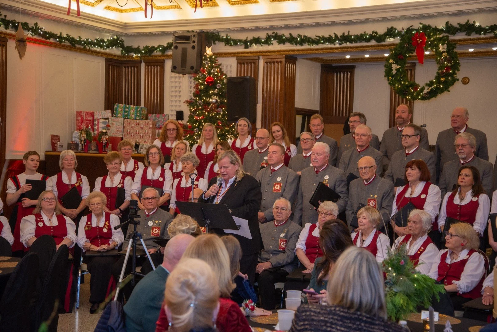 A large choir dressed in traditional holiday attire performing at a Christmas gathering in a decorated hall with a Christmas tree and wreaths.