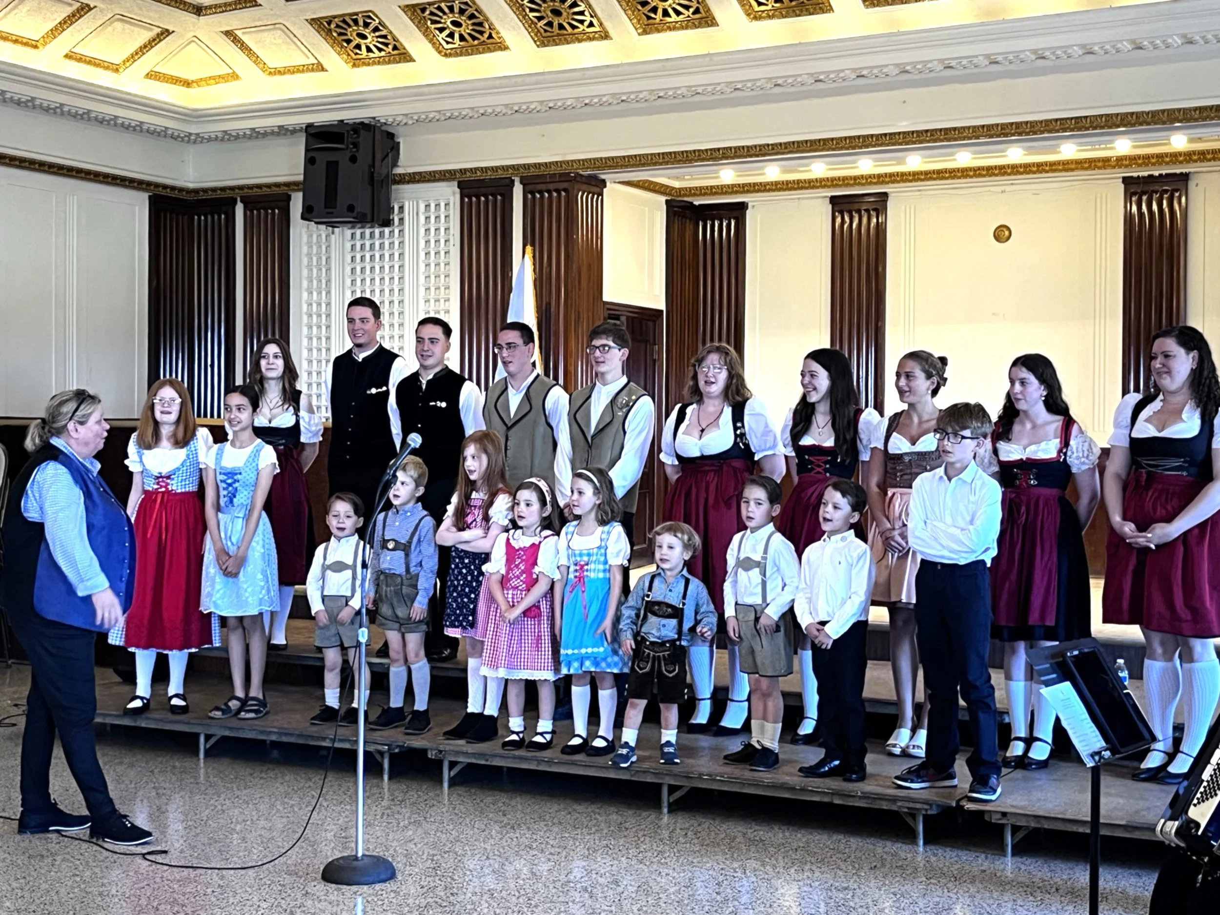 A group of children and adults dressed in traditional Bavarian clothing standing on a stage during a performance or ceremony in a formal room with decorative ceiling and wood paneling.