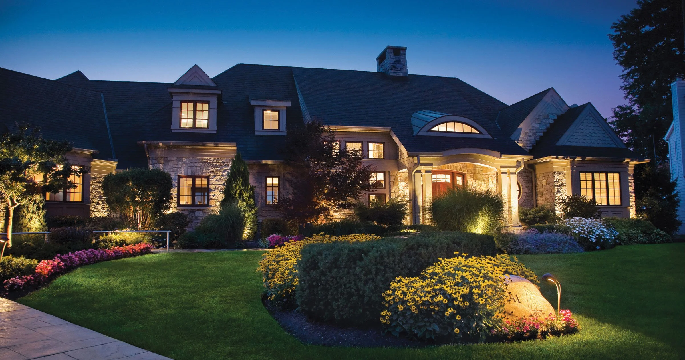 A large, elegant house at dusk with warm lighting. The front yard has a well-maintained lawn, flower beds, shrubs, and pathway lighting, creating a welcoming atmosphere.