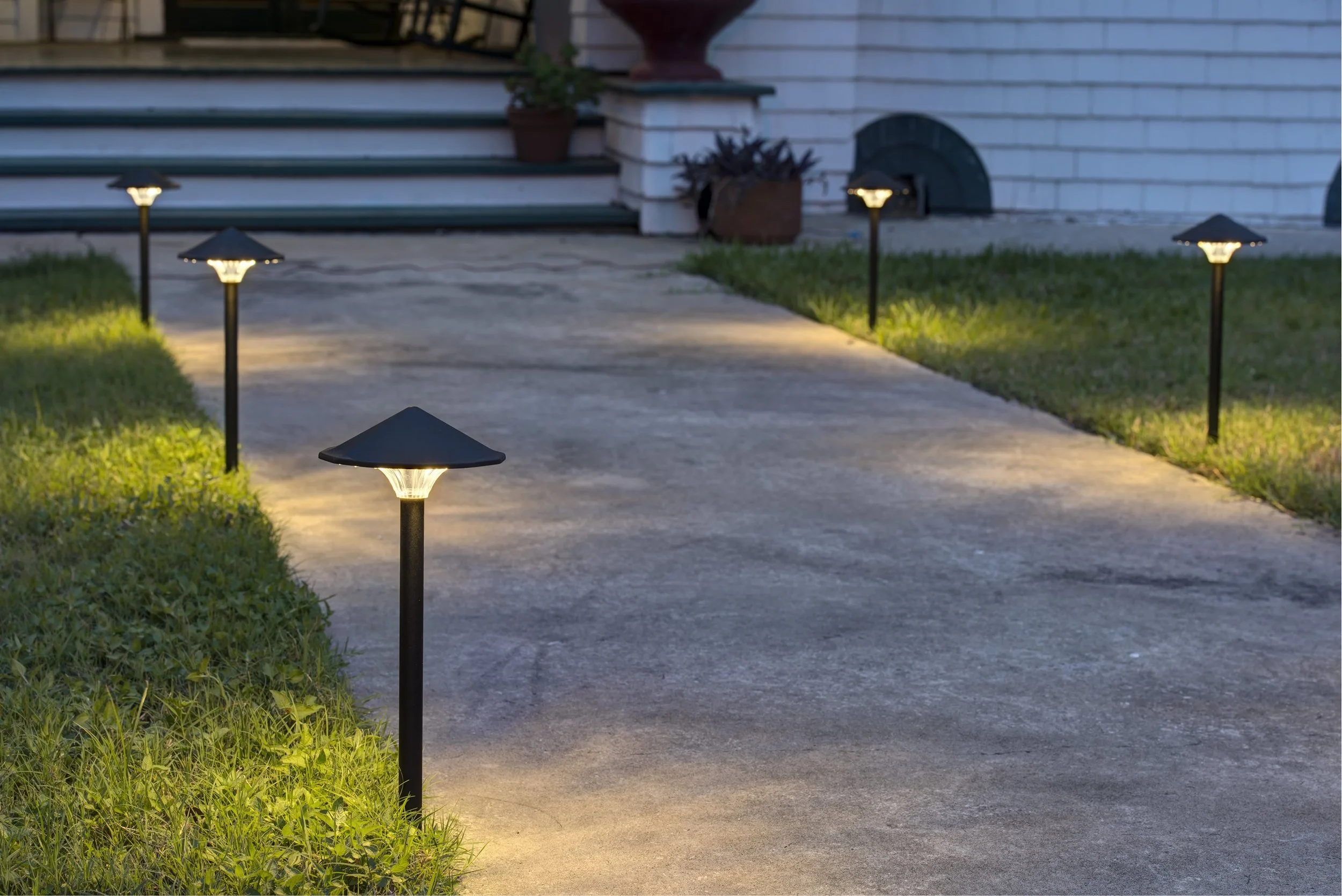 A concrete walkway illuminated by five black garden lights along the edges, leading up to steps of a house with potted plants.