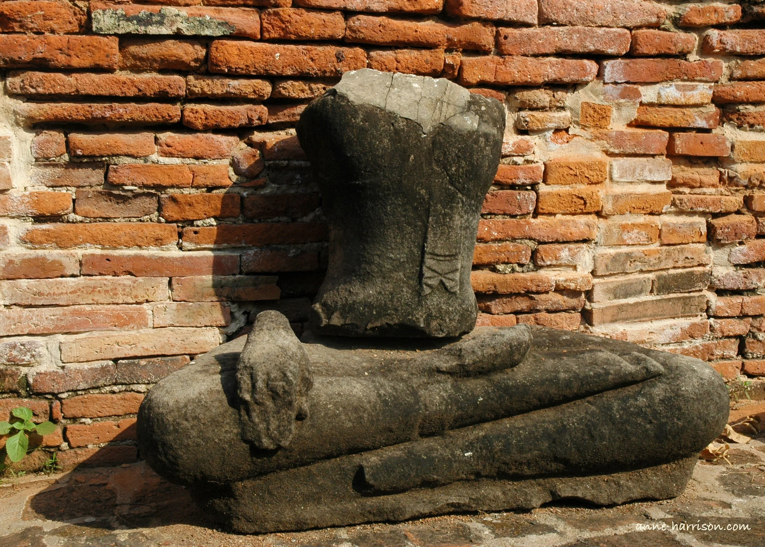 An old statue of a Buddha sits crosslegged in front of a brick wall. He is headless, and lost both arms, although one hand rests on his knee