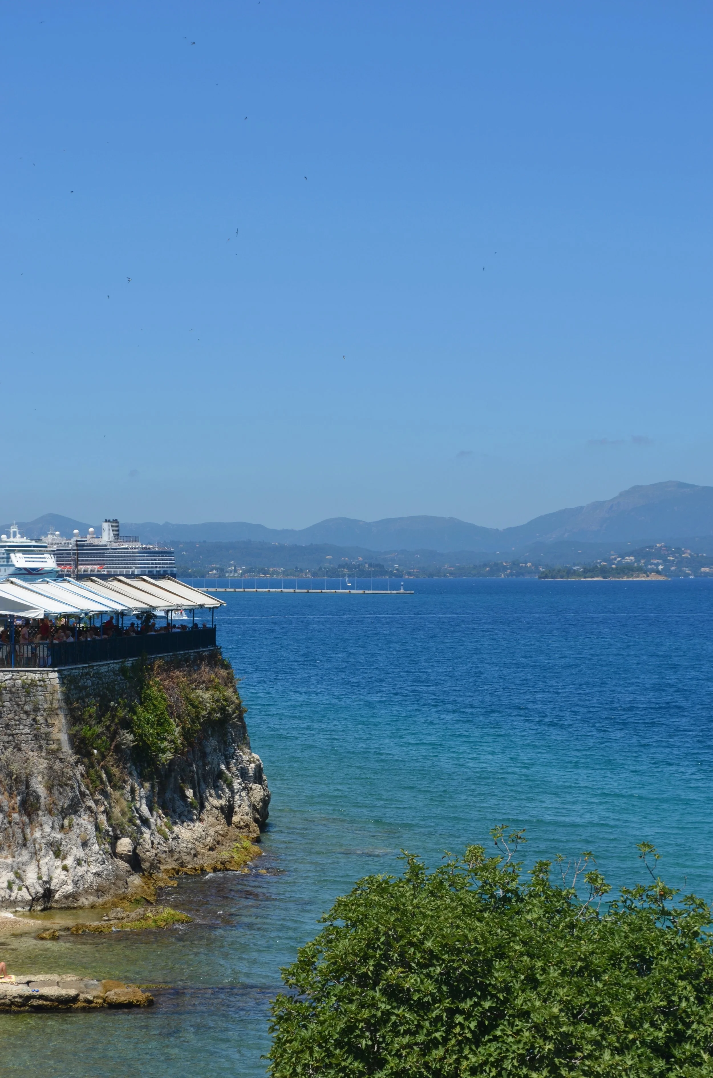 An open-air restaurant covered with shade cloths on a rocky outcrop overlooking Corfu Harbour. The water is blue. A cruise liner is visible in the distance.