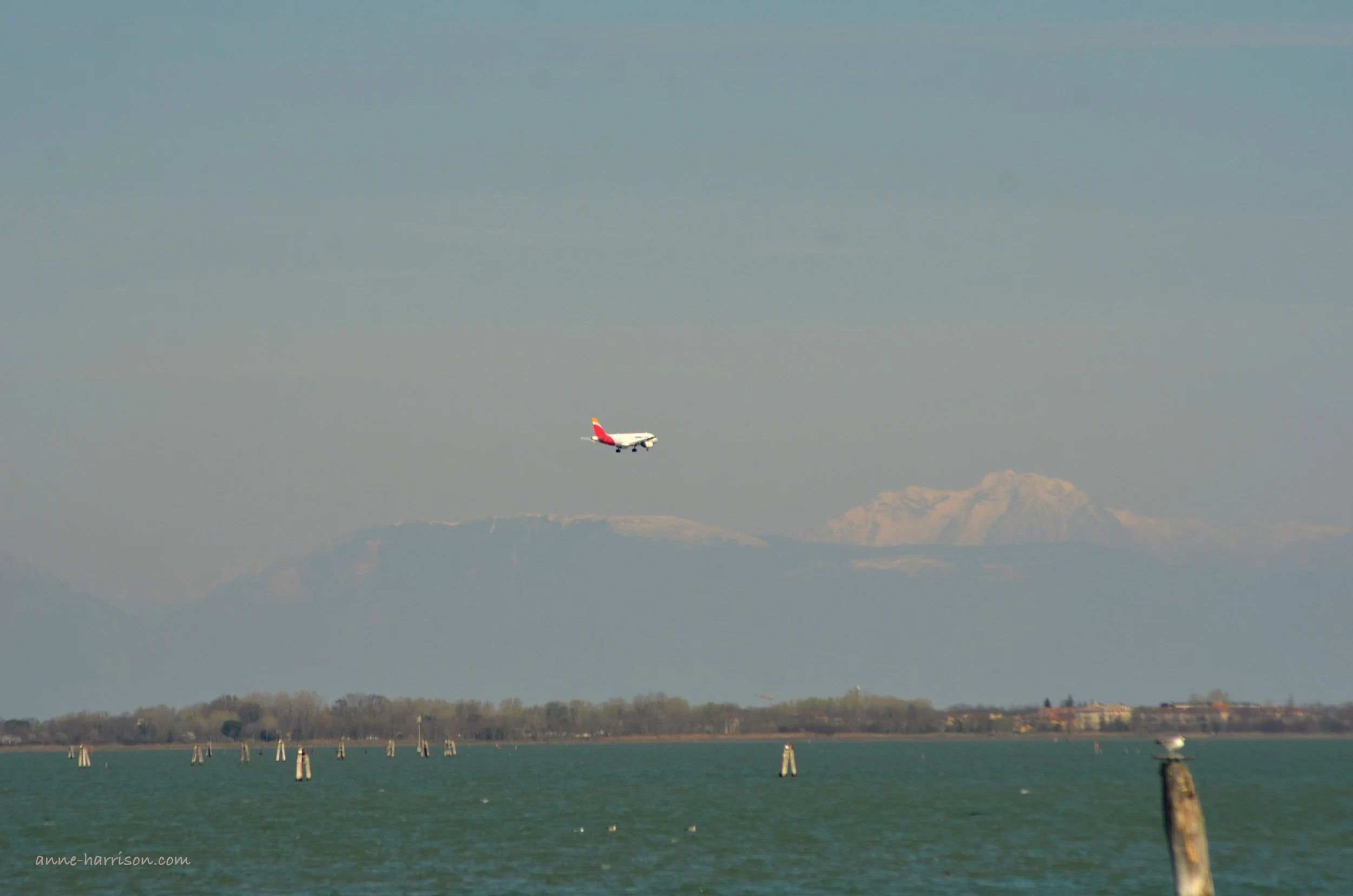A plane is seeing flying over the waters of Venice, with the snow-capped alps in the distance