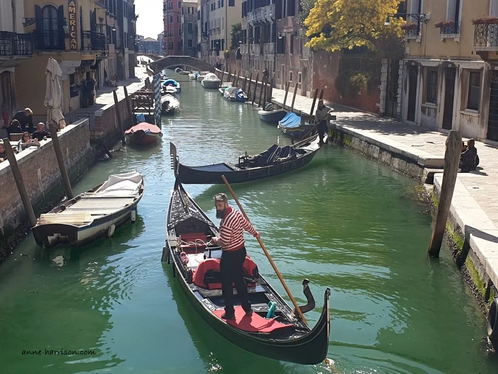 A gondolier in a red stipred shirt steering his gondola down a narrow canal, with another gondola attempting to turn around