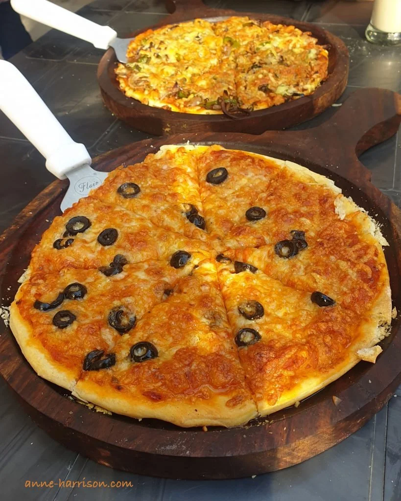 Two pizzas serve don a wooden board. One is covered in olives.