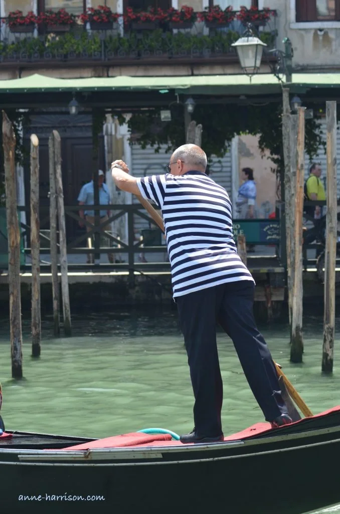 A gondolier steering in his gondola, wearing blue pants and a striped shirt