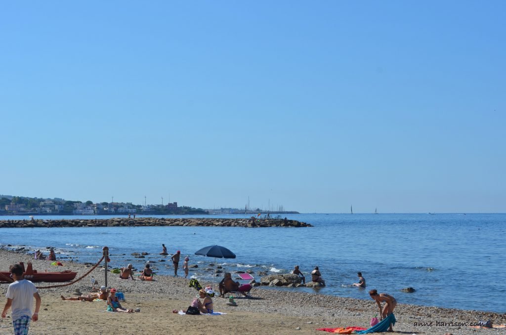 People sun-baking on a stony beach near Rome. The water is blue, and a town is in the distance.