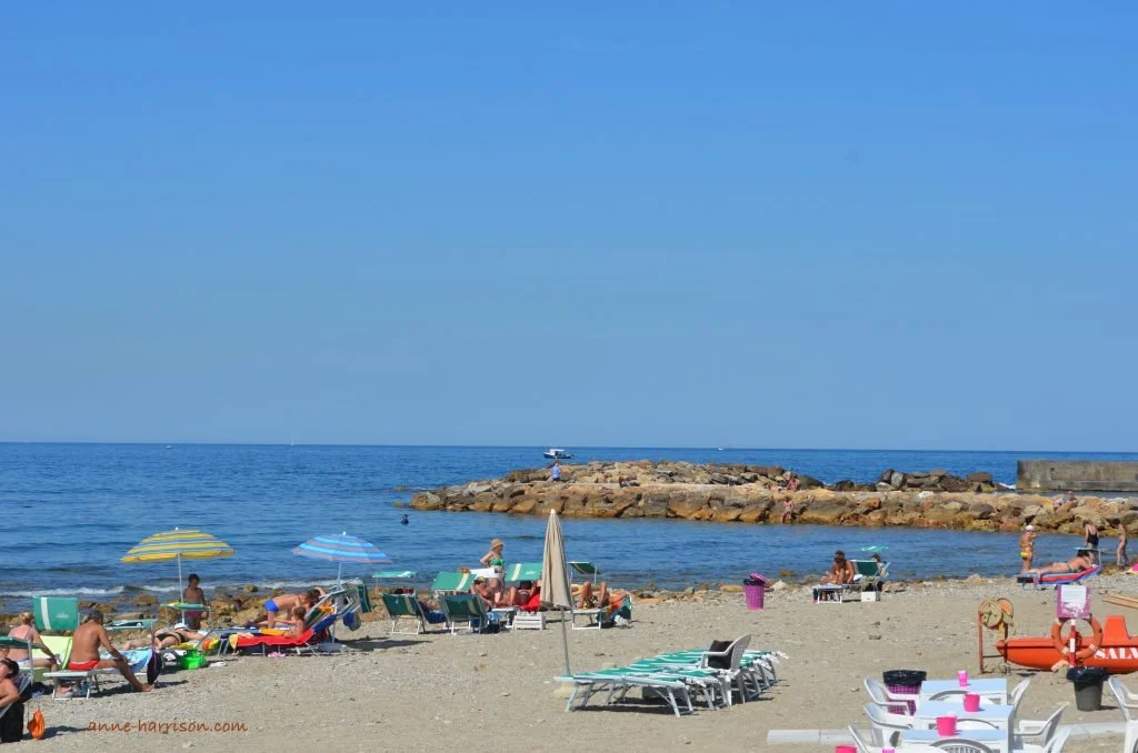 People sunbaking on cahirs on a beach at Civitavecchia, near Rome. The sky is blue, and a break-wall marks the end of the beach.