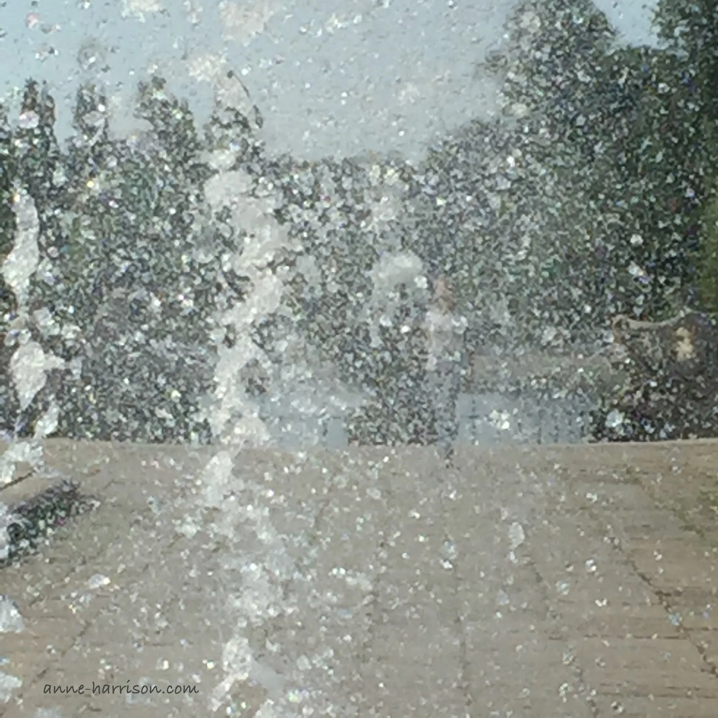 A cloose-up shot of water droplets spraying from a fountain. A tiled footpath and and a pond are just visible.