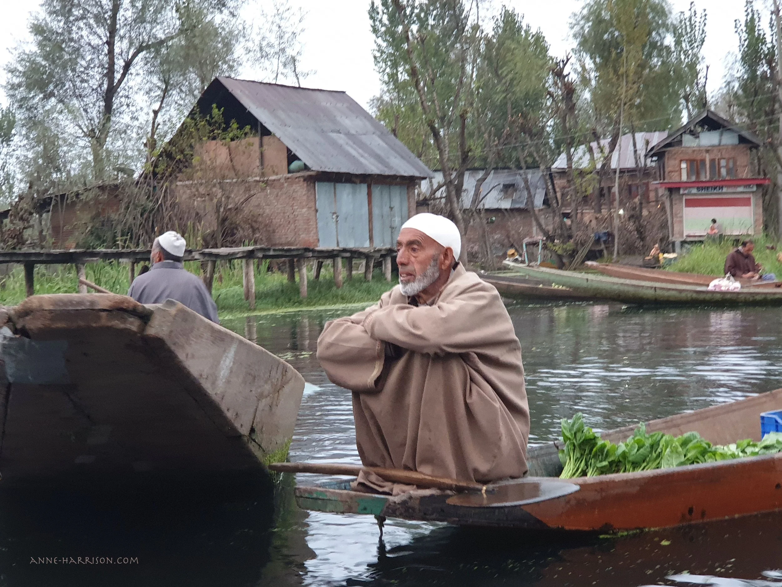An elderly gentleman with a white skull cap sitting on the prow of his wooden boat