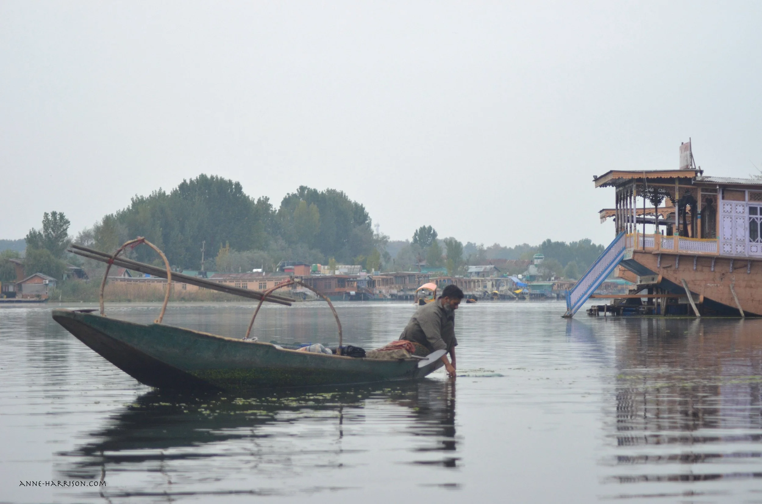 A man in a longboat trailing his hands in the water. Houseboats float on the water.