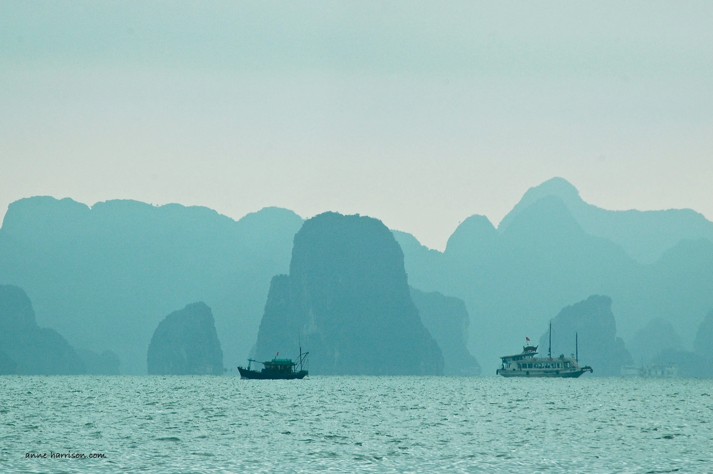 The almost vertical island of Halong Bay, with a fishing boat and tourist boat, at dusk, all in tones of blue.