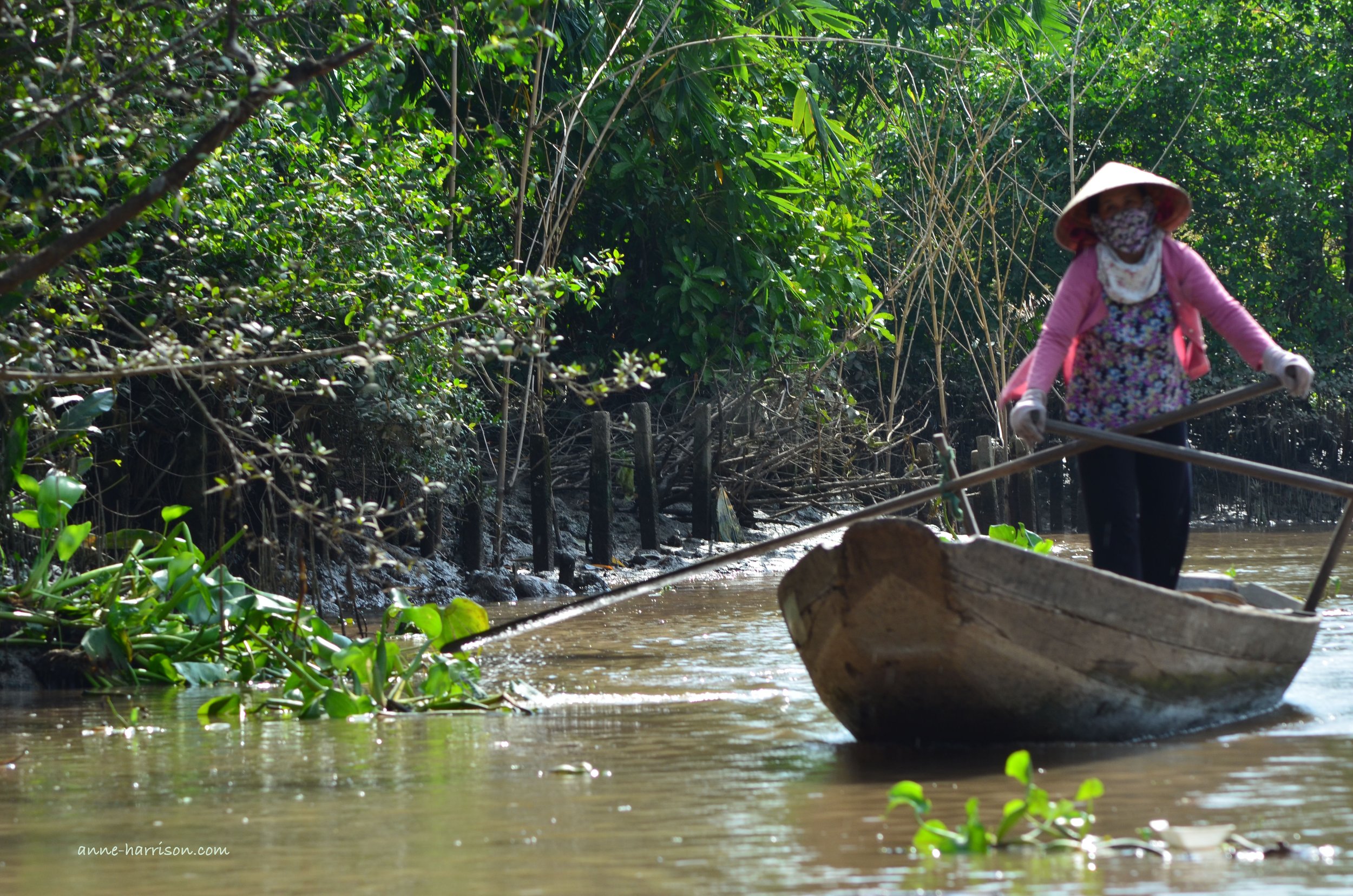 A woman standing on a small wooden row baot. She is facing forward as she rows it. The water is border by lush forest.
